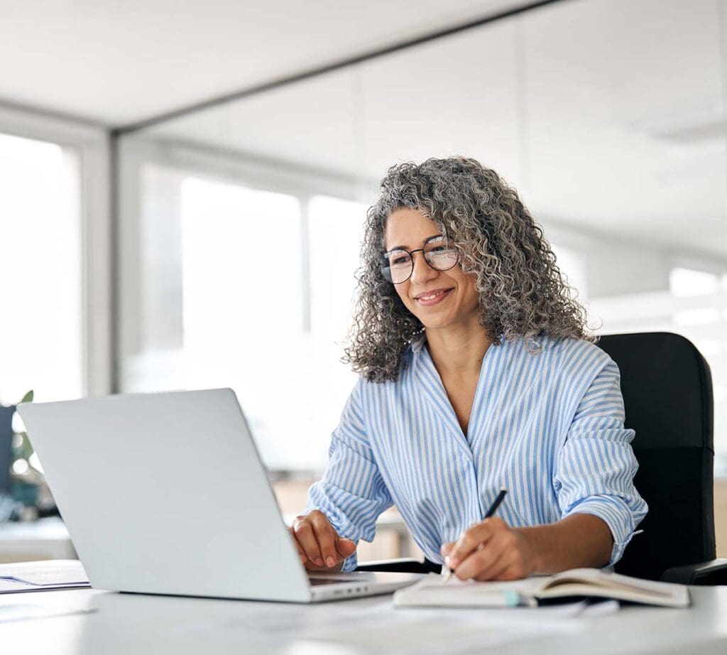 Woman wearing glasses, smiling while working on her laptop.