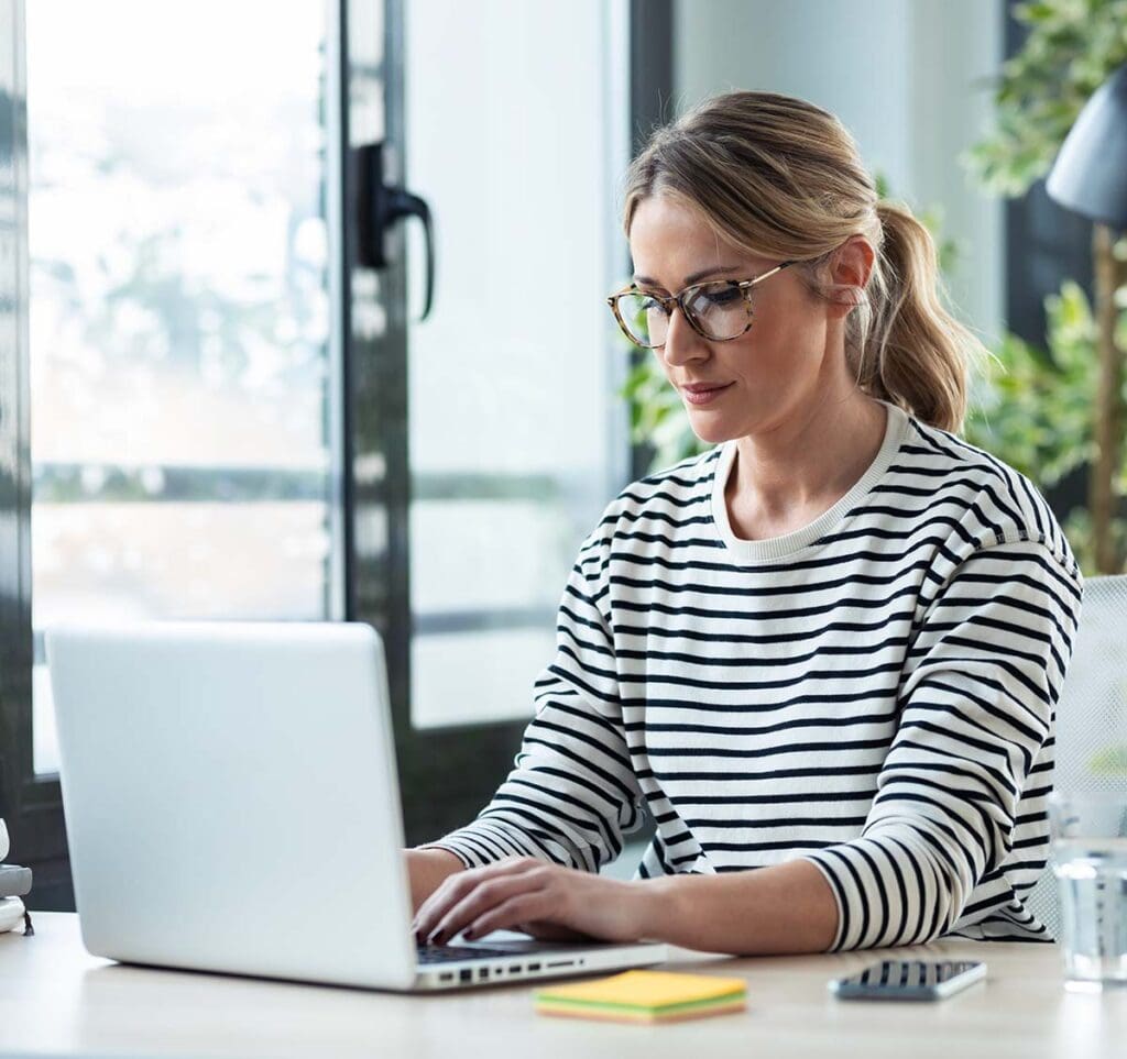 Woman wearing glasses, smiling while working on her laptop.