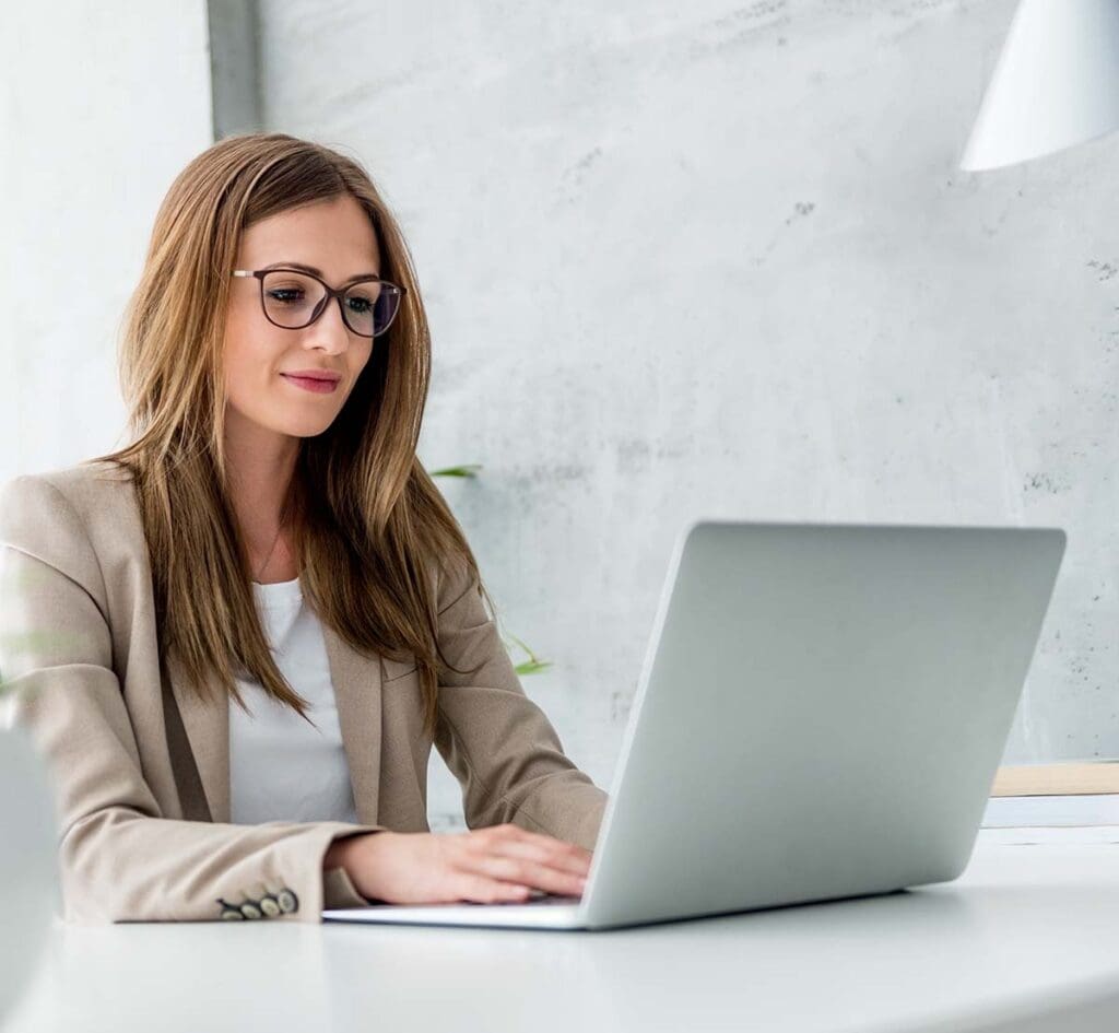 Woman wearing glasses, smiling while working on her laptop.